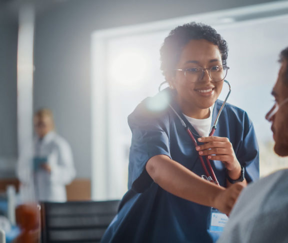 Hospital Ward: Friendly Black Head Nurse Uses Stethoscope to Listen to Heartbeat and Lungs of Recovering Male Patient Resting in Bed, Does Checkup