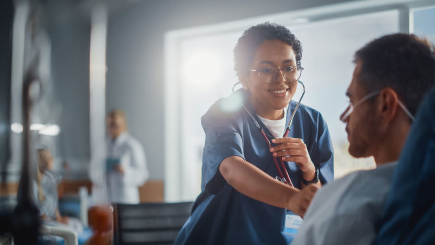 Hospital Ward: Friendly Black Head Nurse Uses Stethoscope to Listen to Heartbeat and Lungs of Recovering Male Patient Resting in Bed, Does Checkup