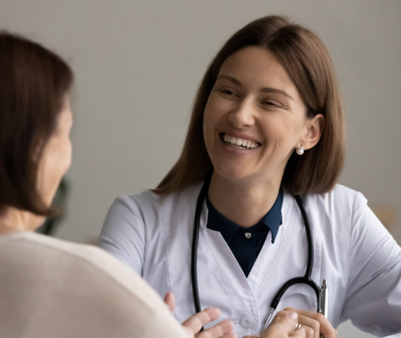 Visiting doctor. Smiling confident female gp attending physician listen to elderly woman visitor on meeting at hospital. Attentive doc feel glad talk to aged woman patient about good treatment results.