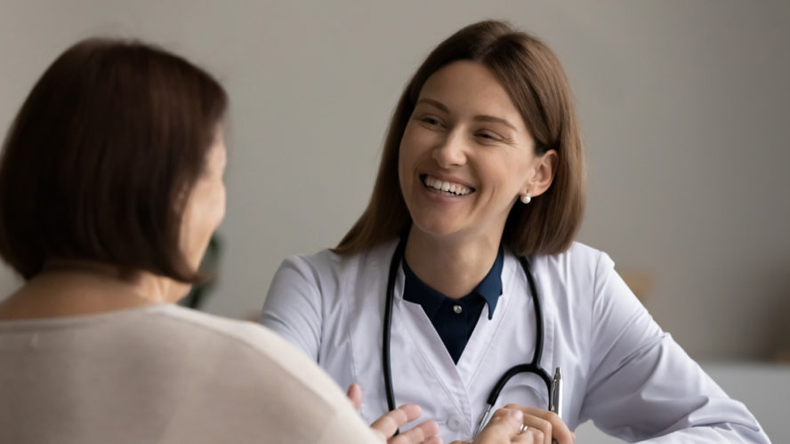 Visiting doctor. Smiling confident female gp attending physician listen to elderly woman visitor on meeting at hospital. Attentive doc feel glad talk to aged woman patient about good treatment results.