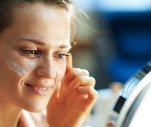 Elegant female applying facial cream and looking in mirror