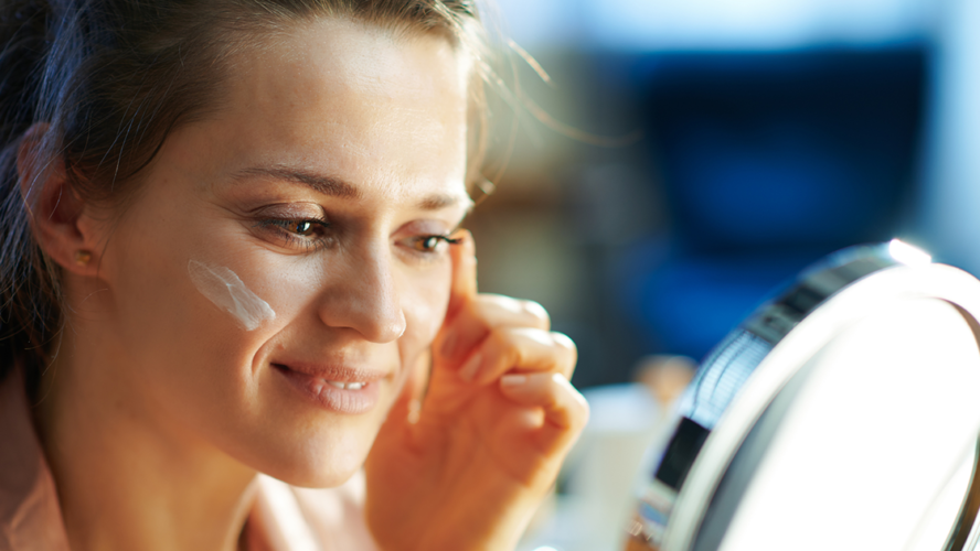 Elegant female applying facial cream and looking in mirror